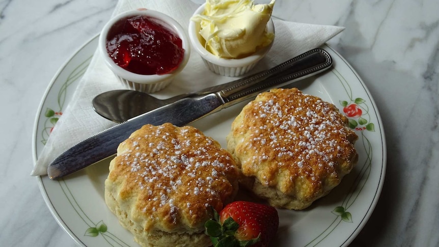 Two scones on a plate, with pots of jam and cream, and a fresh strawberry, beside them.