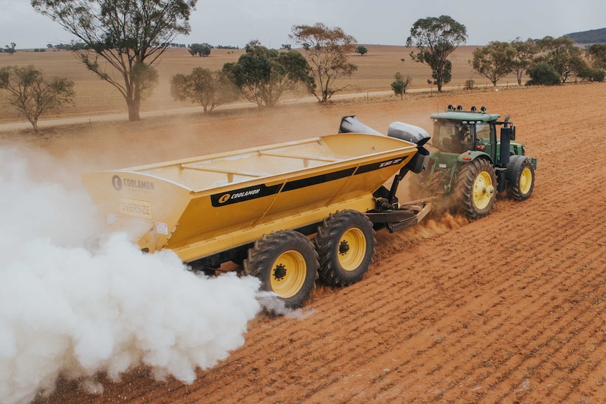 A green tractor in brown stubble towing a yellow spreader chaser bin which is spreading fertiliser for sowing