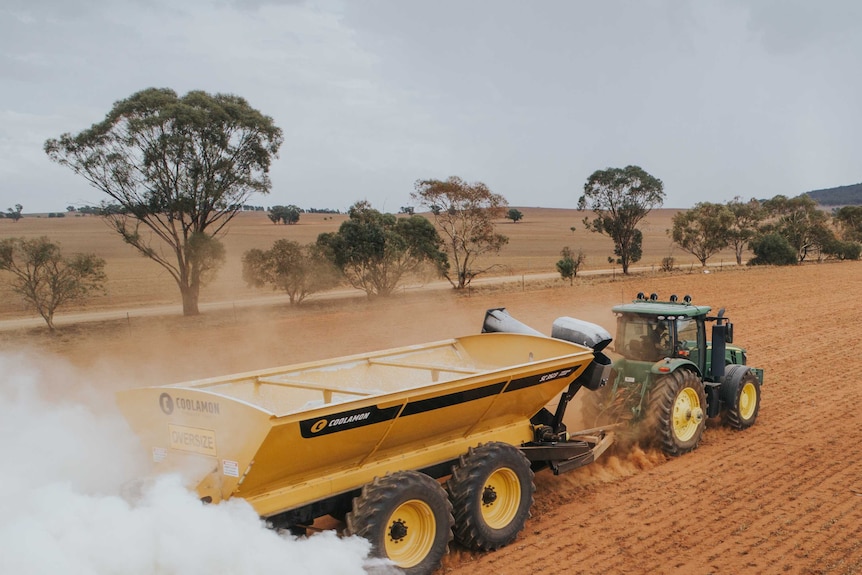 A green tractor in brown stubble towing a yellow spreader chaser bin which is spreading fertiliser for sowing.
