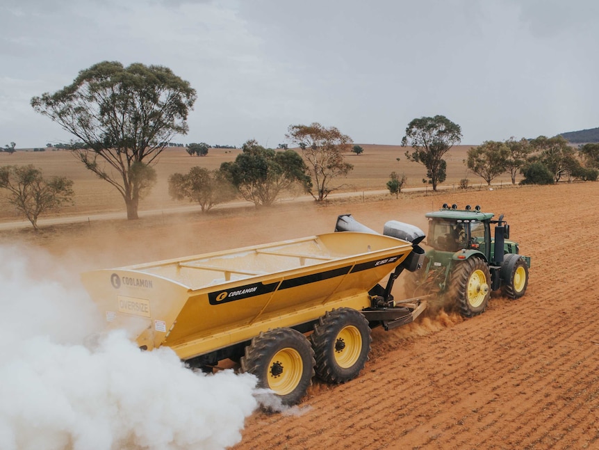 A green tractor in brown stubble towing a yellow chaser bin which is spreading fertiliser for sowing.