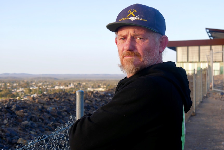 A man in a black hat and jumper stands by a fence and looks straight ahead. 