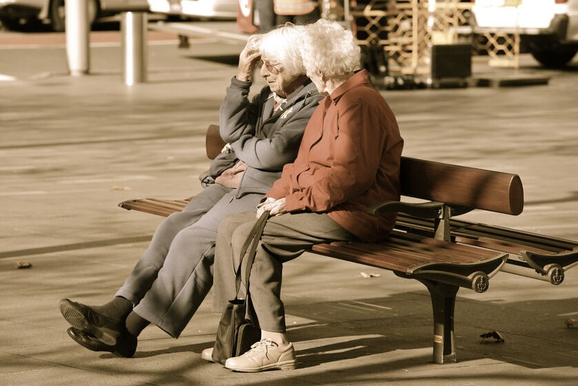 Two women in their 70s sit side-by-side on a bench by a footpath.