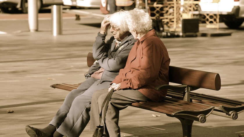 Two women in their 70s sit side-by-side on a bench by a footpath.