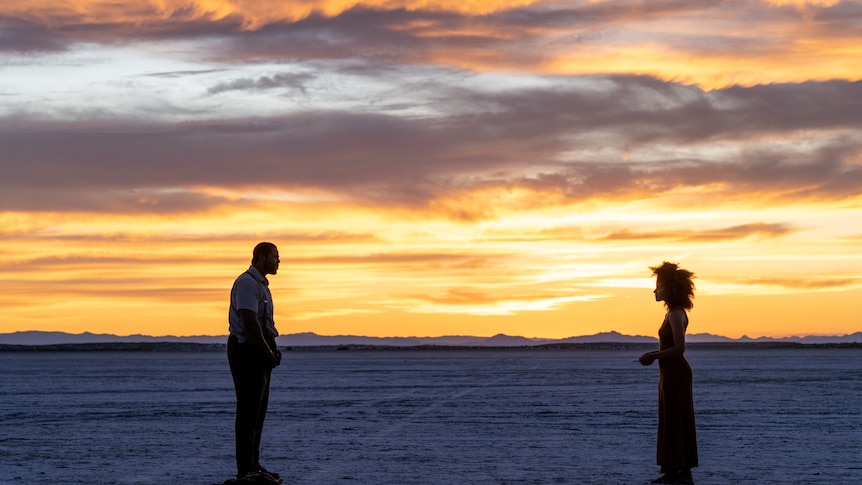 A tall man and a shorter woman stand in a desert as the sun sets, they face each other with a gap between them