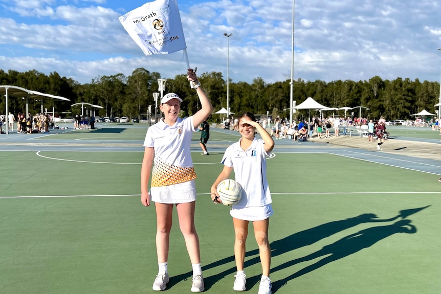 Two young girls wearing white, standing on a netball court. One is holding a white flag above her head.
