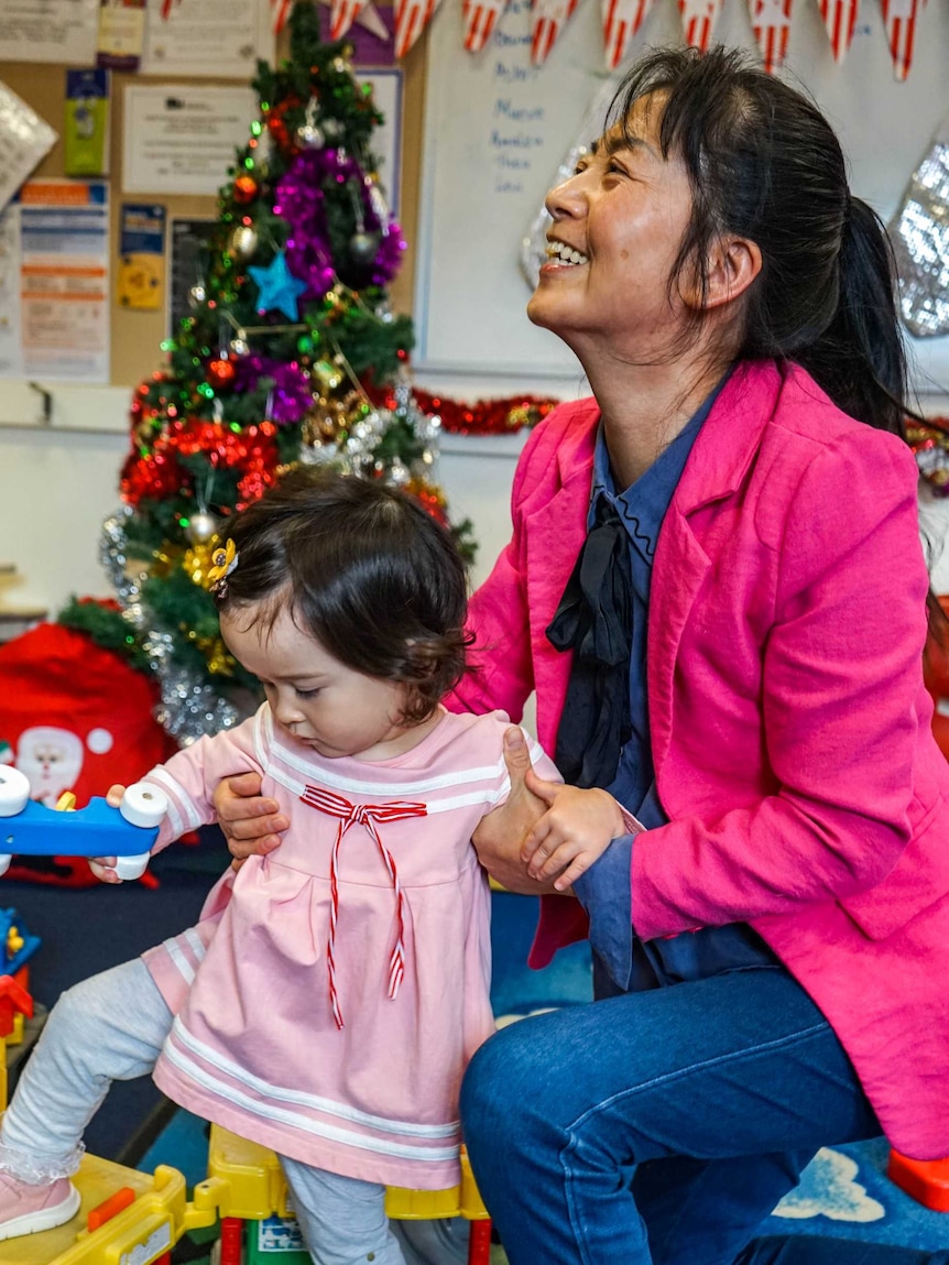 A grandmother kneels beside her granddaughter. A Christmas tree is in the background.