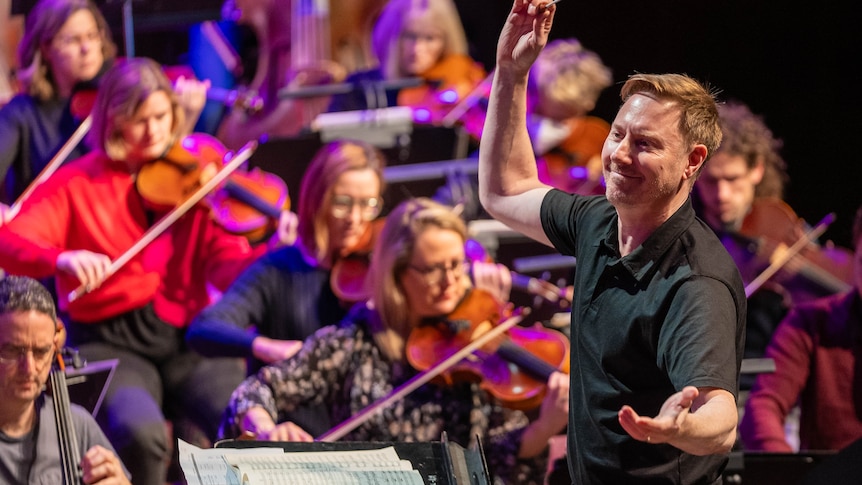 Benjamin Northey conducts the MSO with his baton raised in one hand and his other arm outstretched.