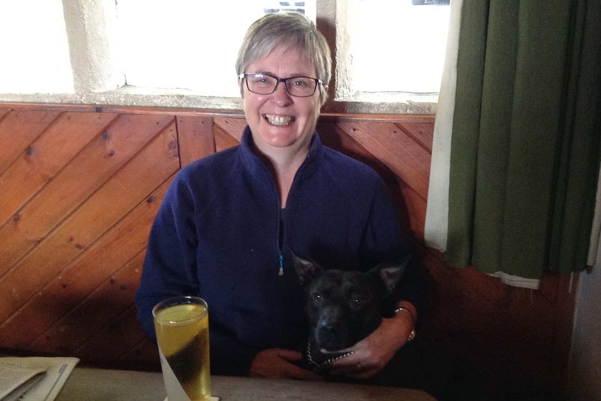 A woman smiles while sitting in a pub cuddling a black dog on her lap.
