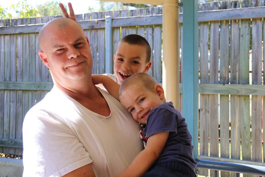 A man holding his two young sons in a backyard