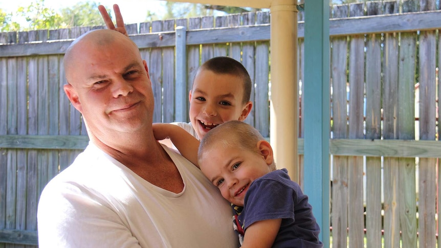 A man holding his two young sons in a backyard