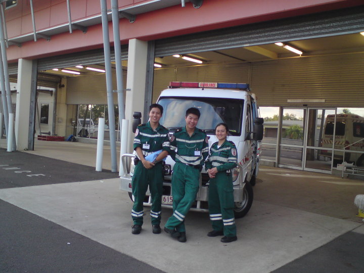 Três pessoas vestidas com uniforme verde de paramédico, em frente a uma ambulância, em uma área de ambulâncias, sorrindo.