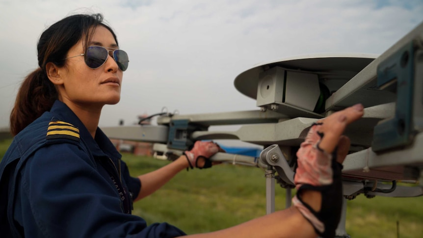 A woman in a pilot's uniform and aviators inspects a chopper's blades