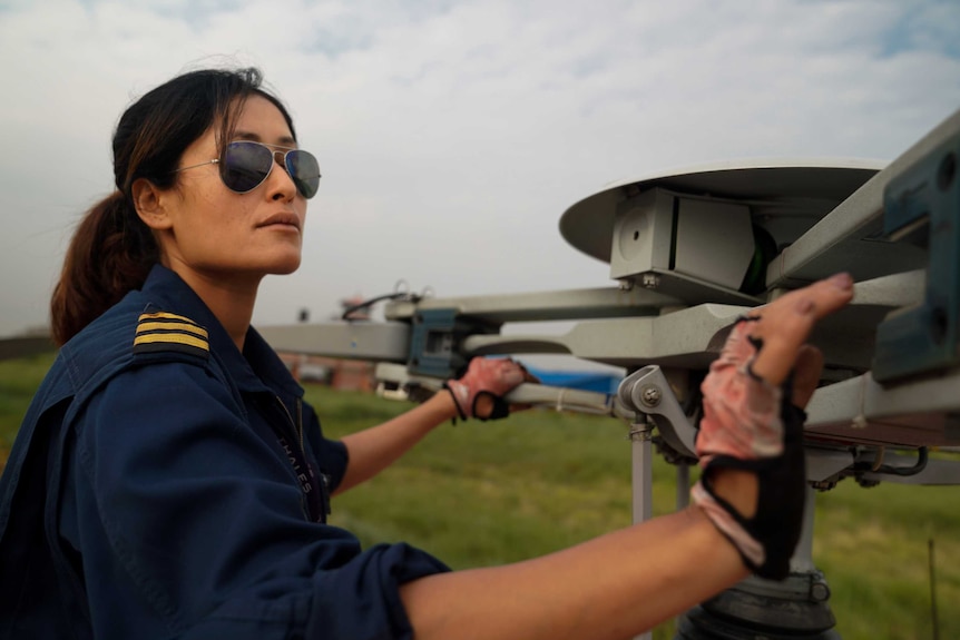 A woman in a pilot's uniform and aviators inspects a chopper's blades