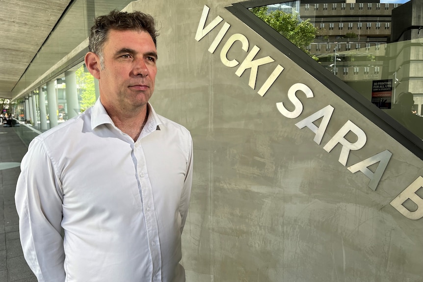 A mid-shot of a man standing in front of a concrete wall looking to his left.