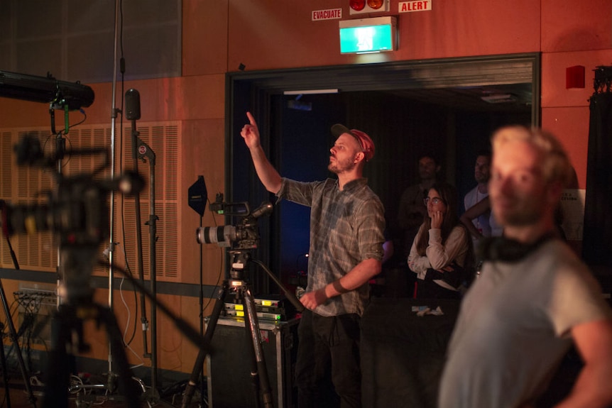 Lavergne stands behind a camera, putting his hand up to say something.