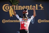 A cyclist wearing a facemask and his white team lycra wears a medal and holds flowers aloft.