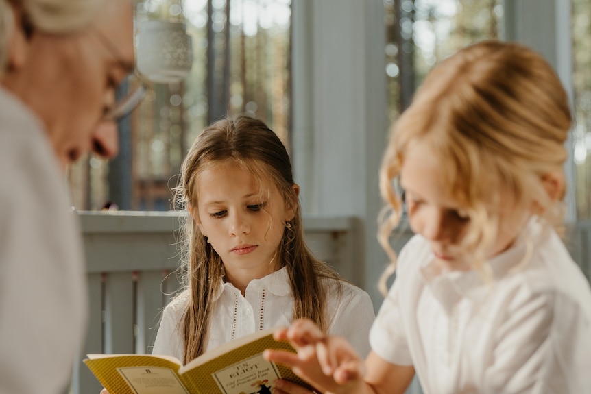 Grandfather playing chess with granddaughter while second granddaughter reads a book.