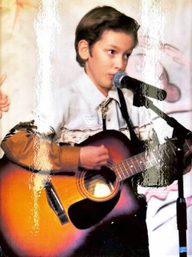 A young boy wears a white shirt and plays the guitar.