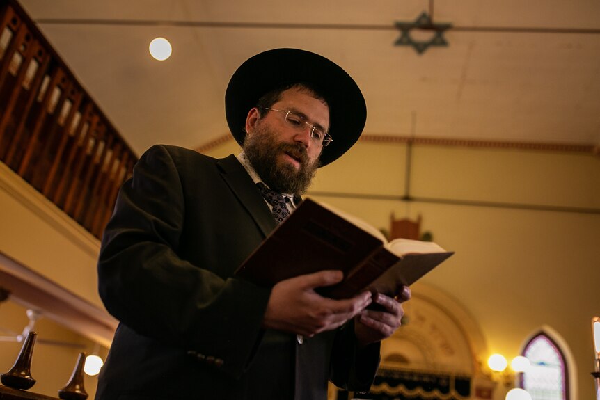 A man wearing a hat reads from a book in a synagogue.