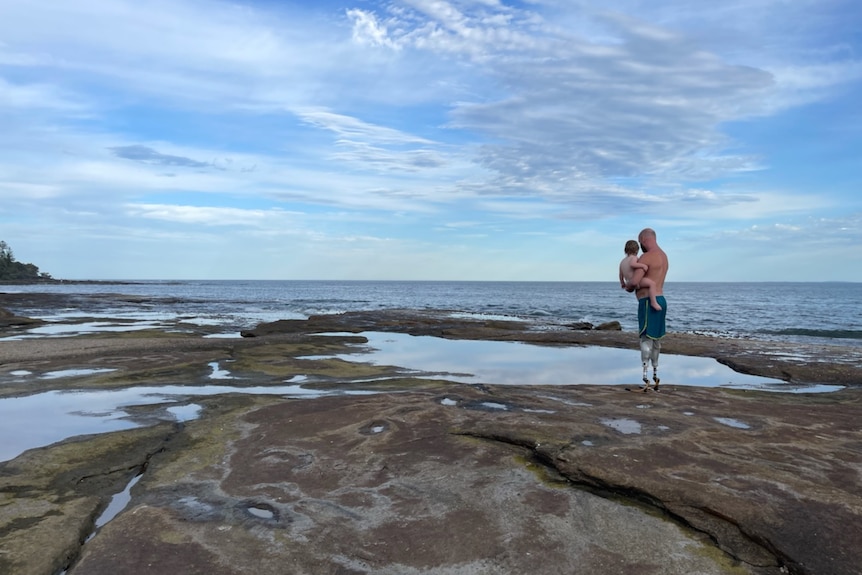 Man and his child standing on the rocks of a beach looking out at the sea, a blue sky with wispy clouds above them