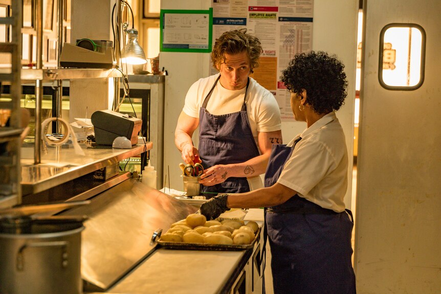 A young white man and an older Latina both in a white tshirt and blue apron prepare food in a commercial kitchen