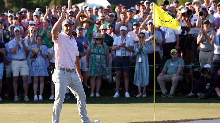 An American golfer stands on the 18th green at the Masters holding his ball in the air as the crowd cheers.