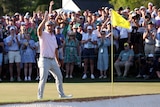 An American golfer stands on the 18th green at the Masters holding his ball in the air as the crowd cheers.