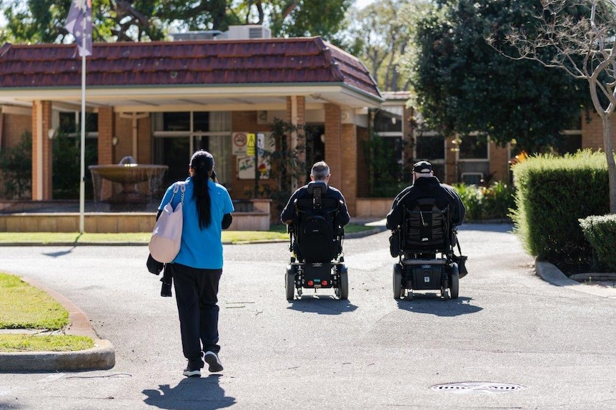 Two men in wheelchairs in front of a building with a healthcare worker in a blue shirt.