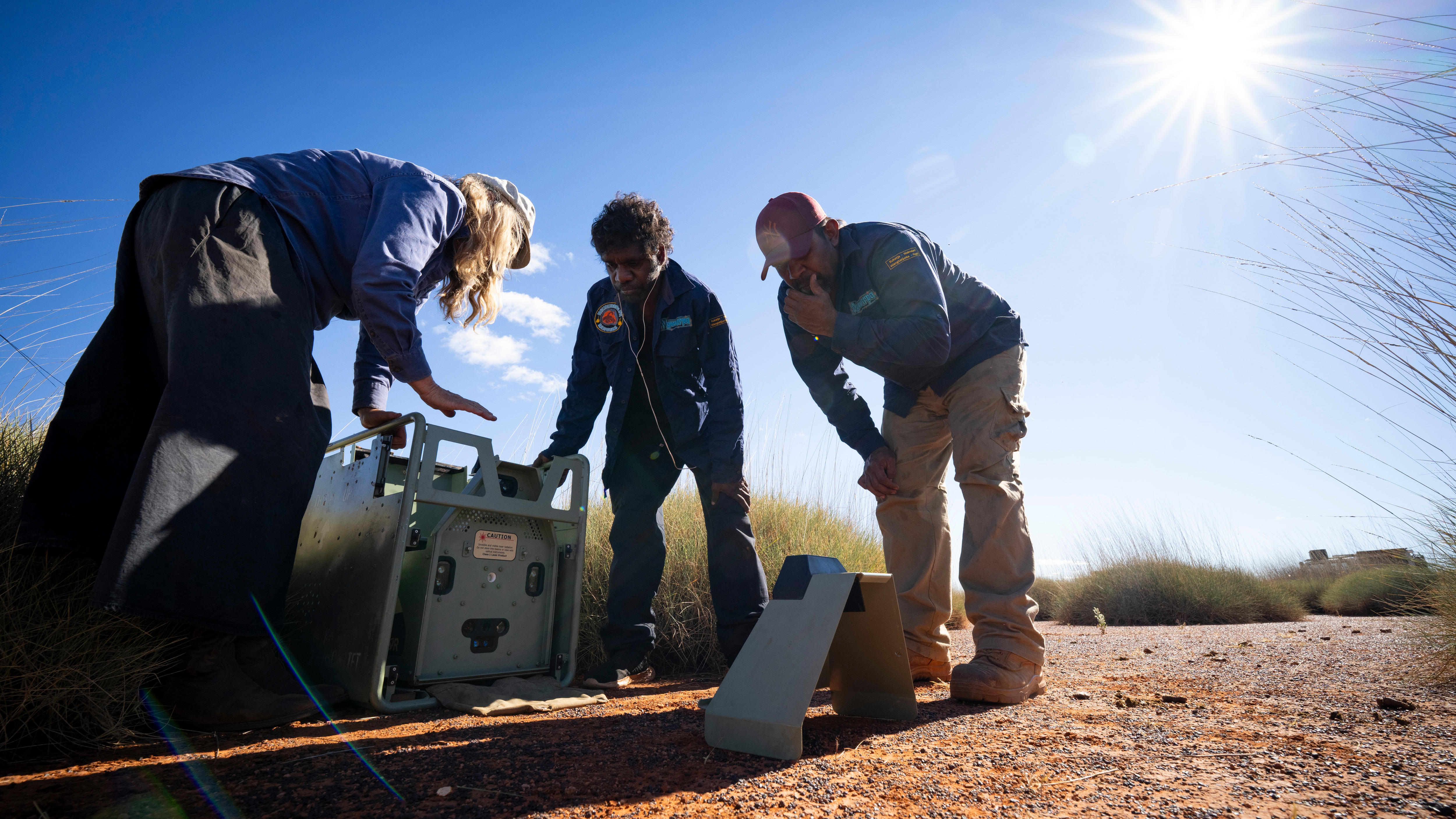 Three people crouched over a box-shaped device on a orange hard ground near low clumps of spinifex.