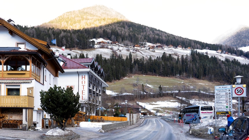 A street bends past hotels leading up into mountains which have snow and trees on them.