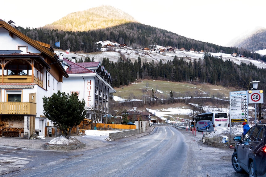 A street bends past hotels leading up into mountains which have snow and trees on them.