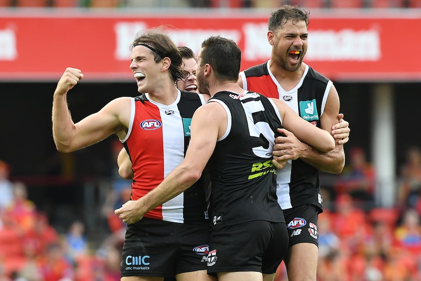 An AFL player pumps his fist in joy after scoring a goal as he is surrounded by teammates.