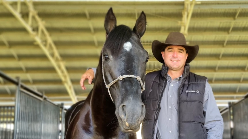 A man in a brown hat and black vest stands beside a black horse.
