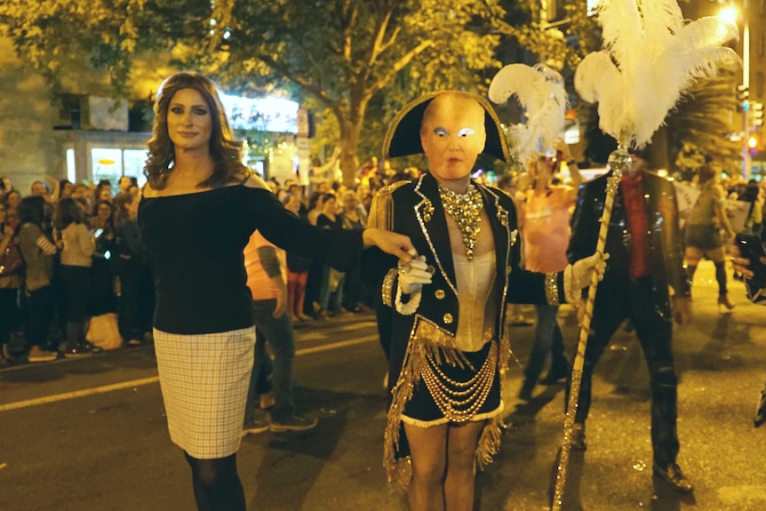 Drag queens dressed as Donald Trump and his wife, Melania, standing on a street at night-time, looked upon by spectators