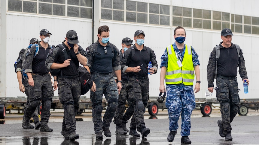 Men in fatigues and wearing masks walk along wet ground