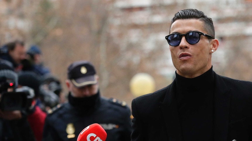 Cristiano Ronaldo in sunglasses and a black trench coat arrives at a Madrid court as a reporter holds out a red microphone.