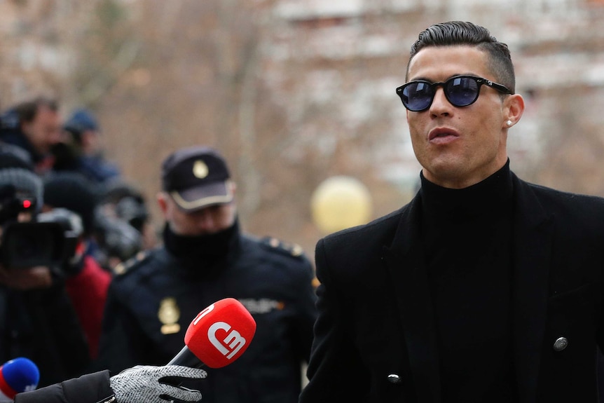 Cristiano Ronaldo in sunglasses and a black trench coat arrives at a Madrid court as a reporter holds out a red microphone.