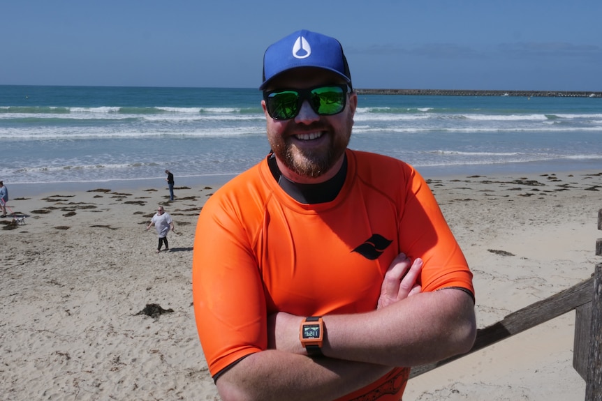 A man wearing orange rashie and cap and sunglasses grins on beach