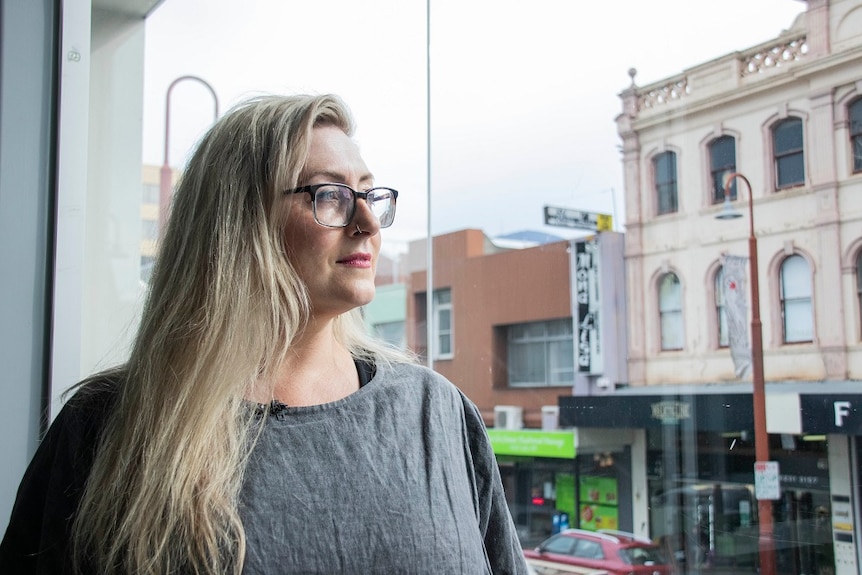 A blonde woman with glasses solemnly looks out over a street scene from a balcony on the first floor.