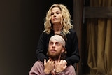 Sigrid Thornton in a blonde wig onstage stands behind a seated Harry Greenwood wearing a bandage on his head.