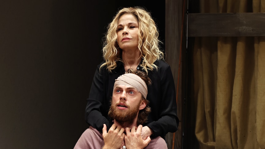 Sigrid Thornton in a blonde wig onstage stands behind a seated Harry Greenwood wearing a bandage on his head.