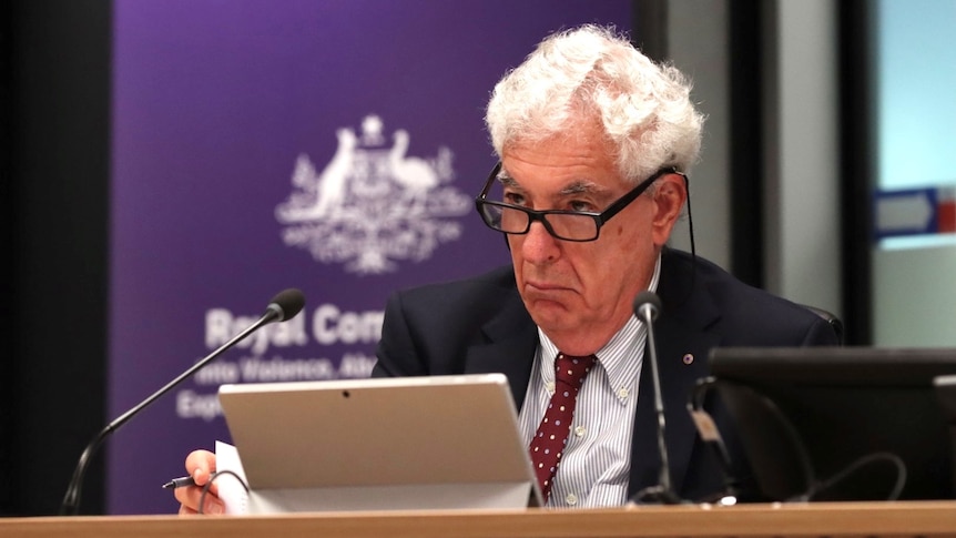 A man with grey hair looks seriously over the top of his glasses.