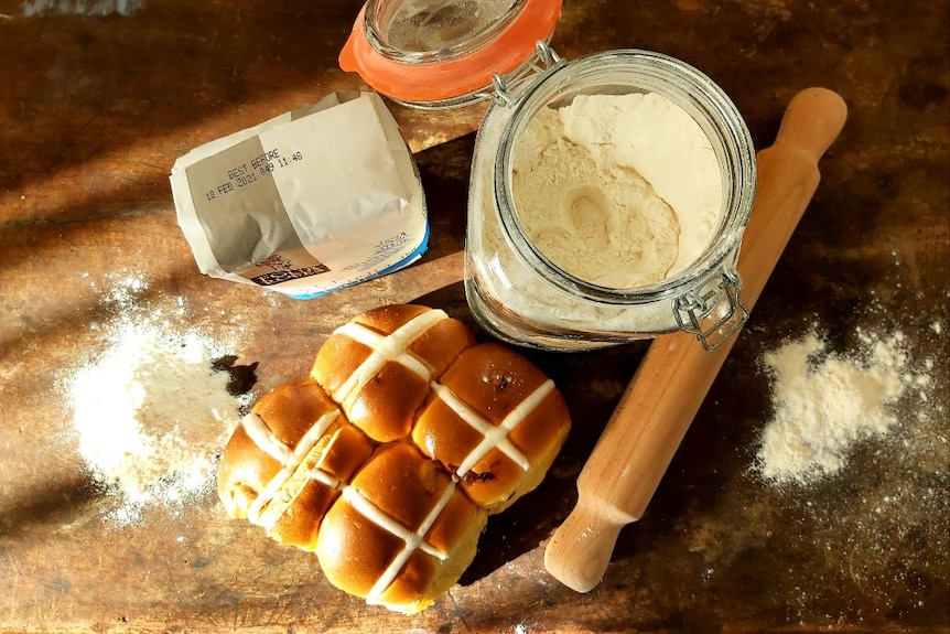 An open jar of flour, a packet of flour, a rolling pin and hot cross buns are arranged on a table