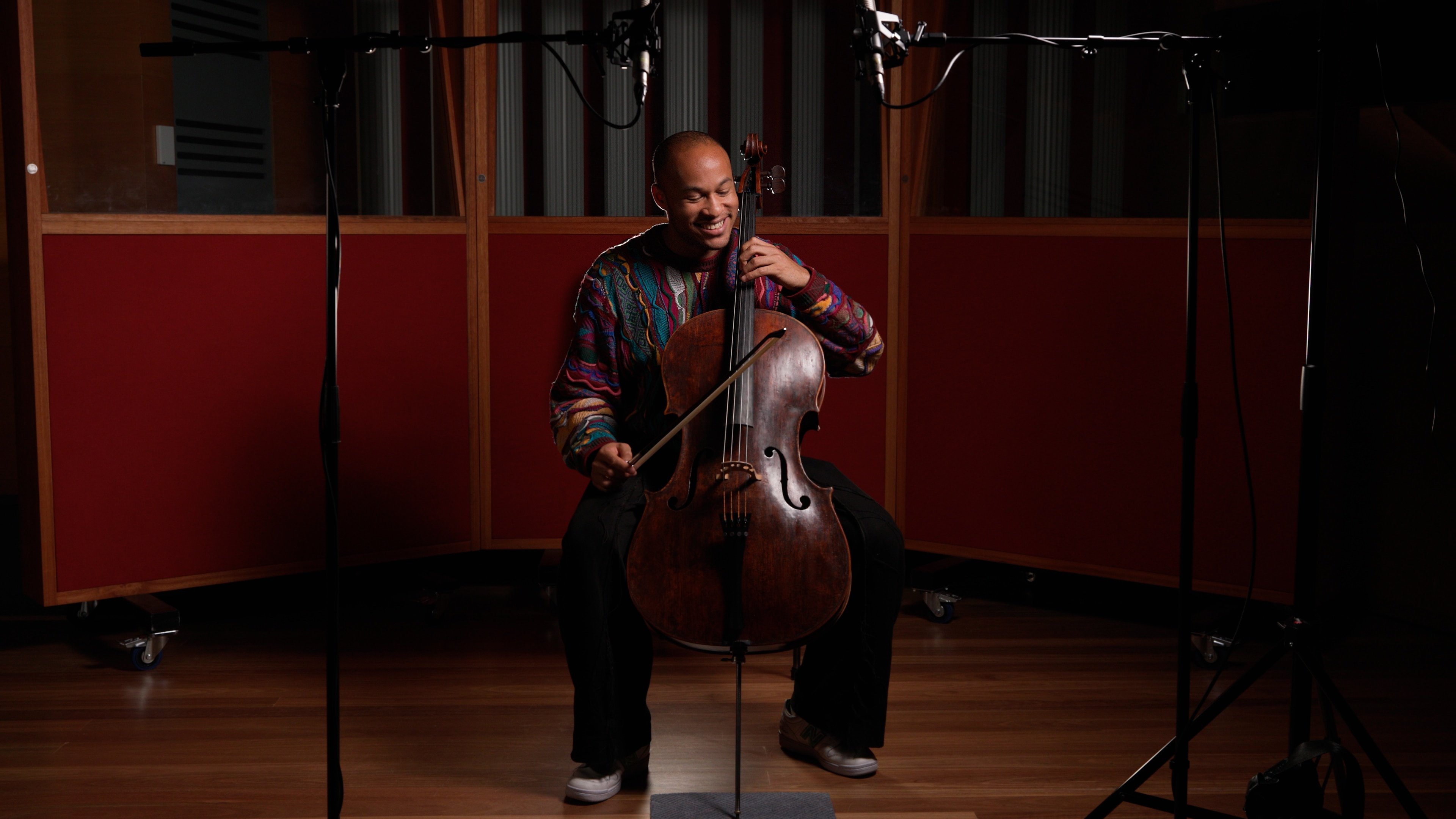 Sheku Kanneh-Mason wears a bright colourful jumper, sitting with his cello ready to play in a music studio with a wooden floor