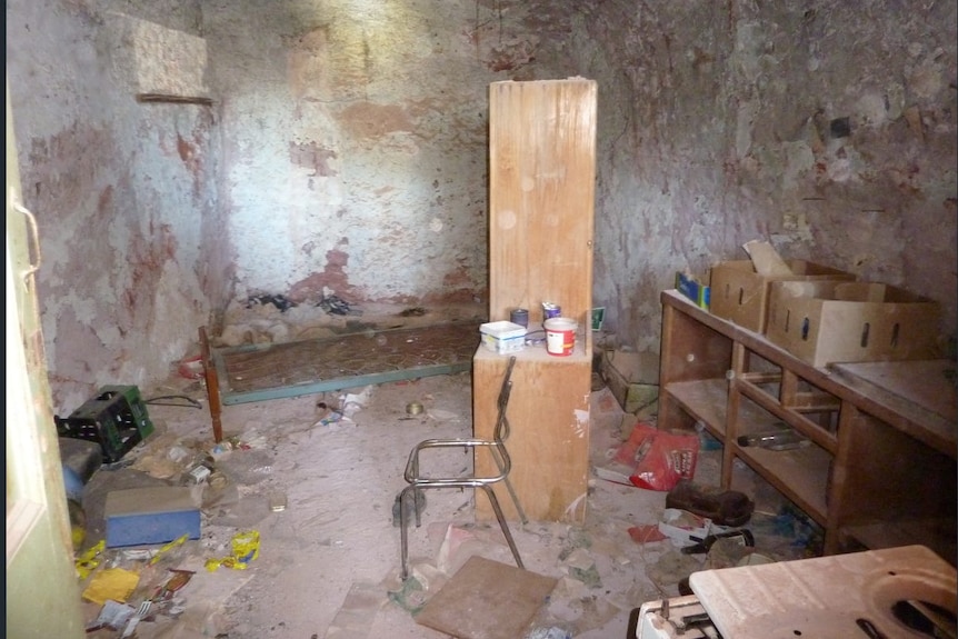 An interior shot of a vacant dugout with a dusty chair, cupboard, shelves and debris on the ground