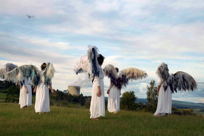 A group of people with angel wings and white robes stand on a green hill, power station in the background