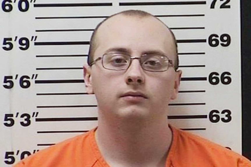 Jake Patterson wears an orange prison-issued jumpsuit and glasses as he stares into the camera against measuring backdrop.