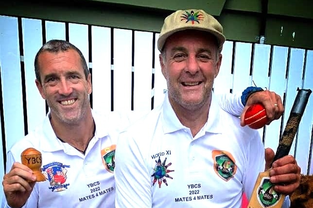 A man in a white cricket shirt holding the Ashes and cricket ball next to another man wearing a cricket cap holding a bat.