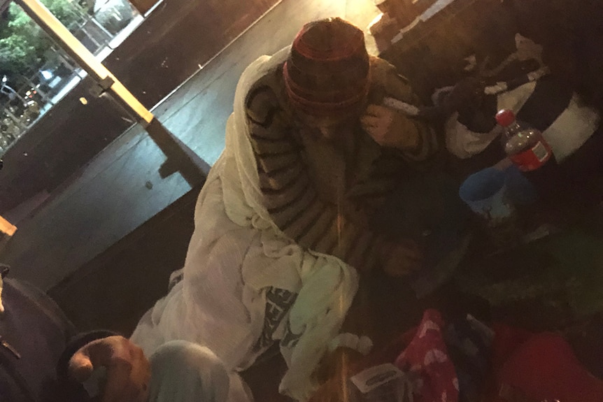 A man wearing a sweater and beanie laying under a blanket on a footpath in Brisbane CBD at night.