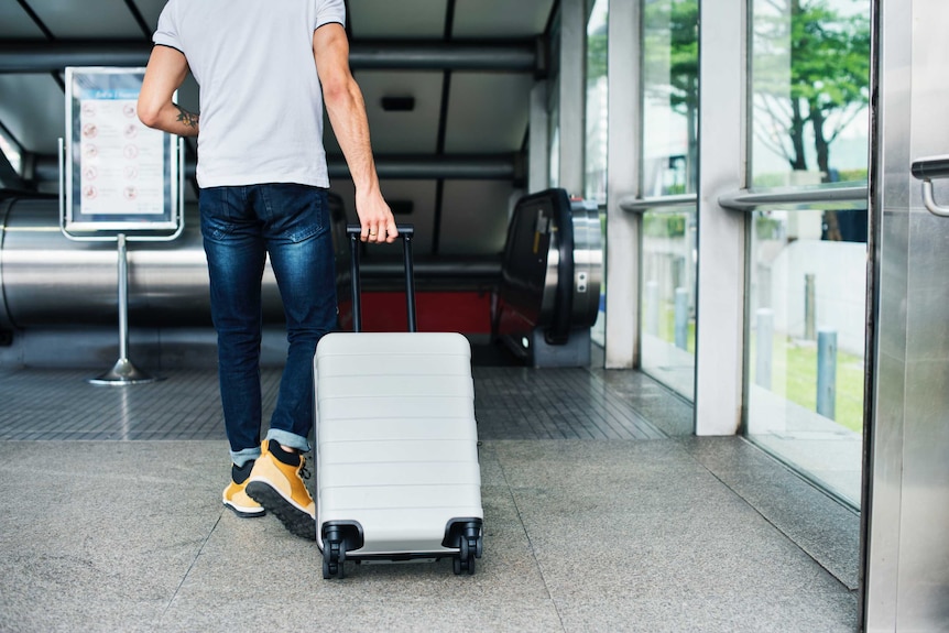 A young man wheels his carry on luggage through an airport, towards an escalator.